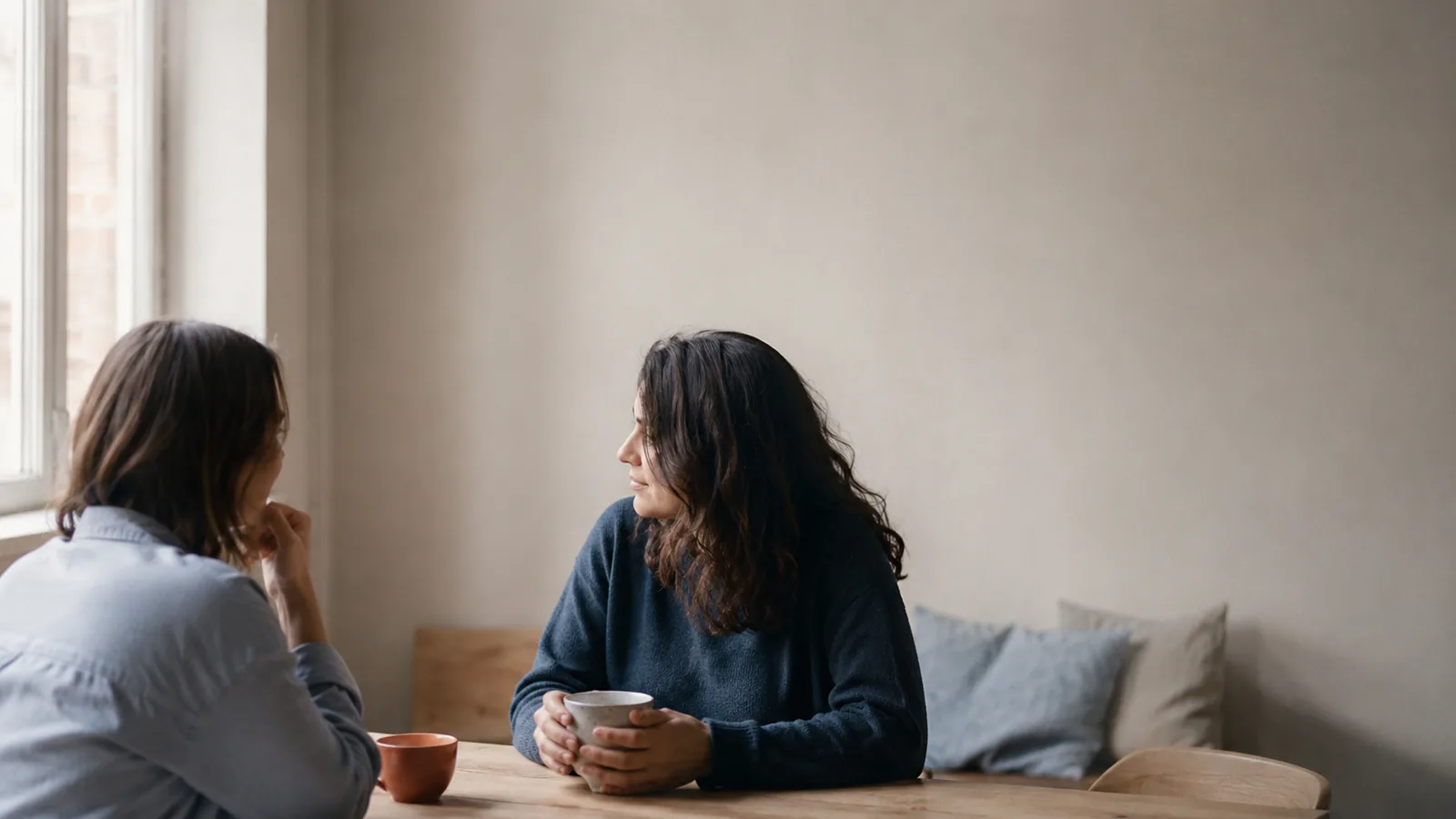 Duas mulheres numa conversa tranquila junto a uma janela, com chávenas sobre uma mesa de madeira.